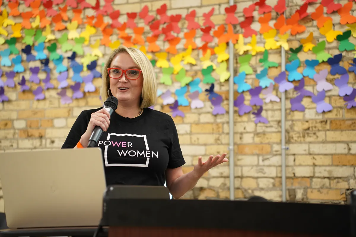 A speaker at the podium in a Power Women shirt at ARKAST 2025
