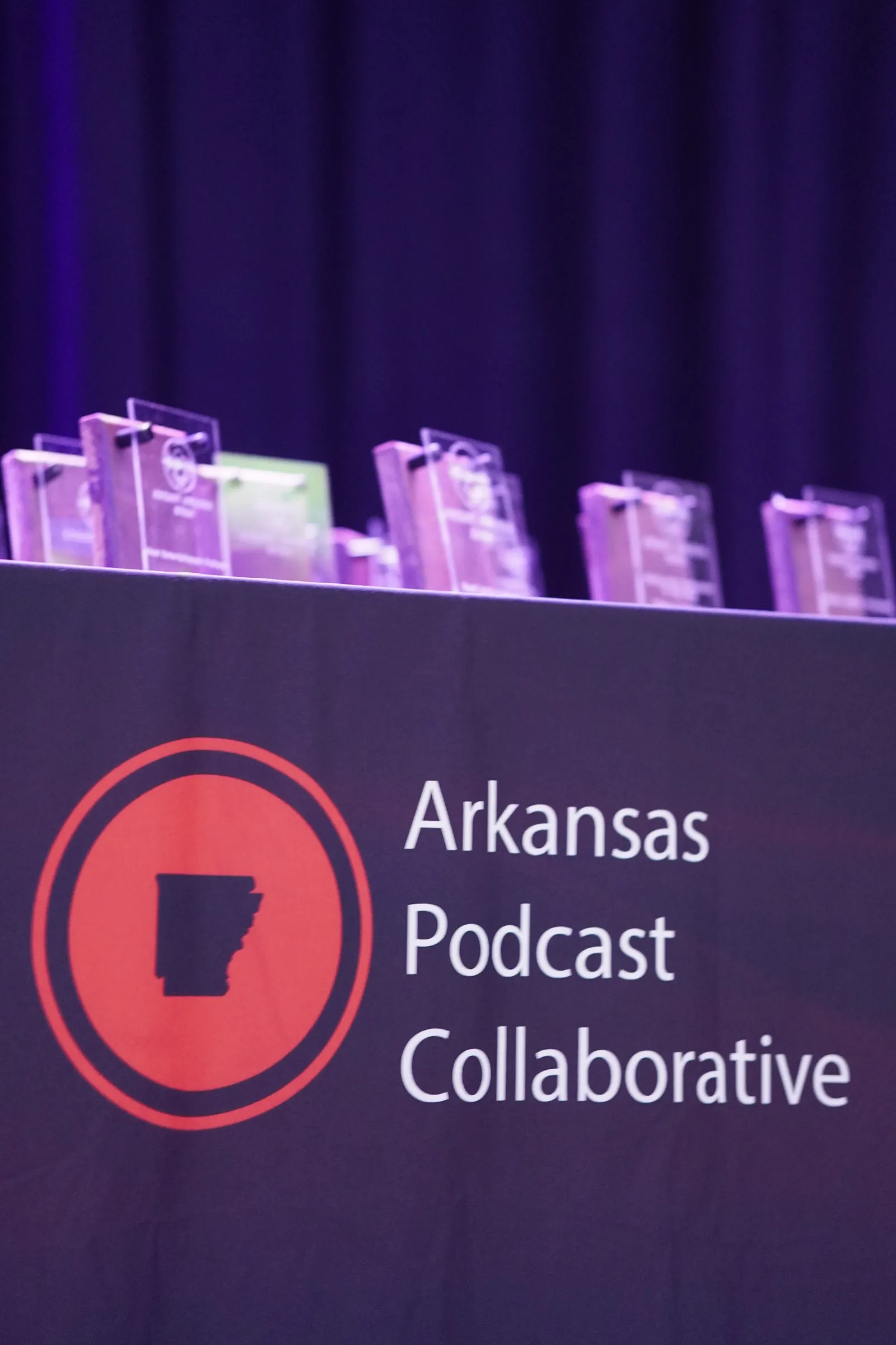 Acrylic ARKAST Awards trophies lined up on a table behind an Arkansas Podcast Collaborative banner